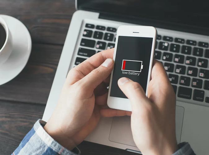 ​Mężczyzna  siedzący przy biurku i trzymający telefon w dłoni, gdzie na ekranie telefonu widać wskaźnik rozładowanej baterii.